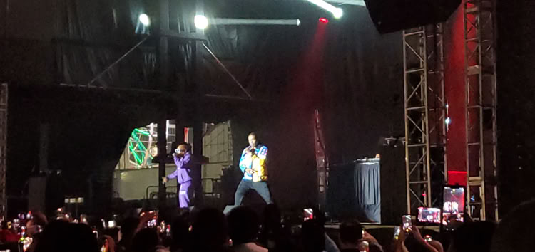 Two performers on stage with microphones, surrounded by stage lights and equipment, in front of an audience holding up phones.