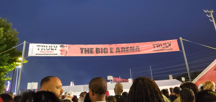 A crowd of people standing under a large banner that reads "The Big E Arena" at night, with a tent and trees visible in the background.